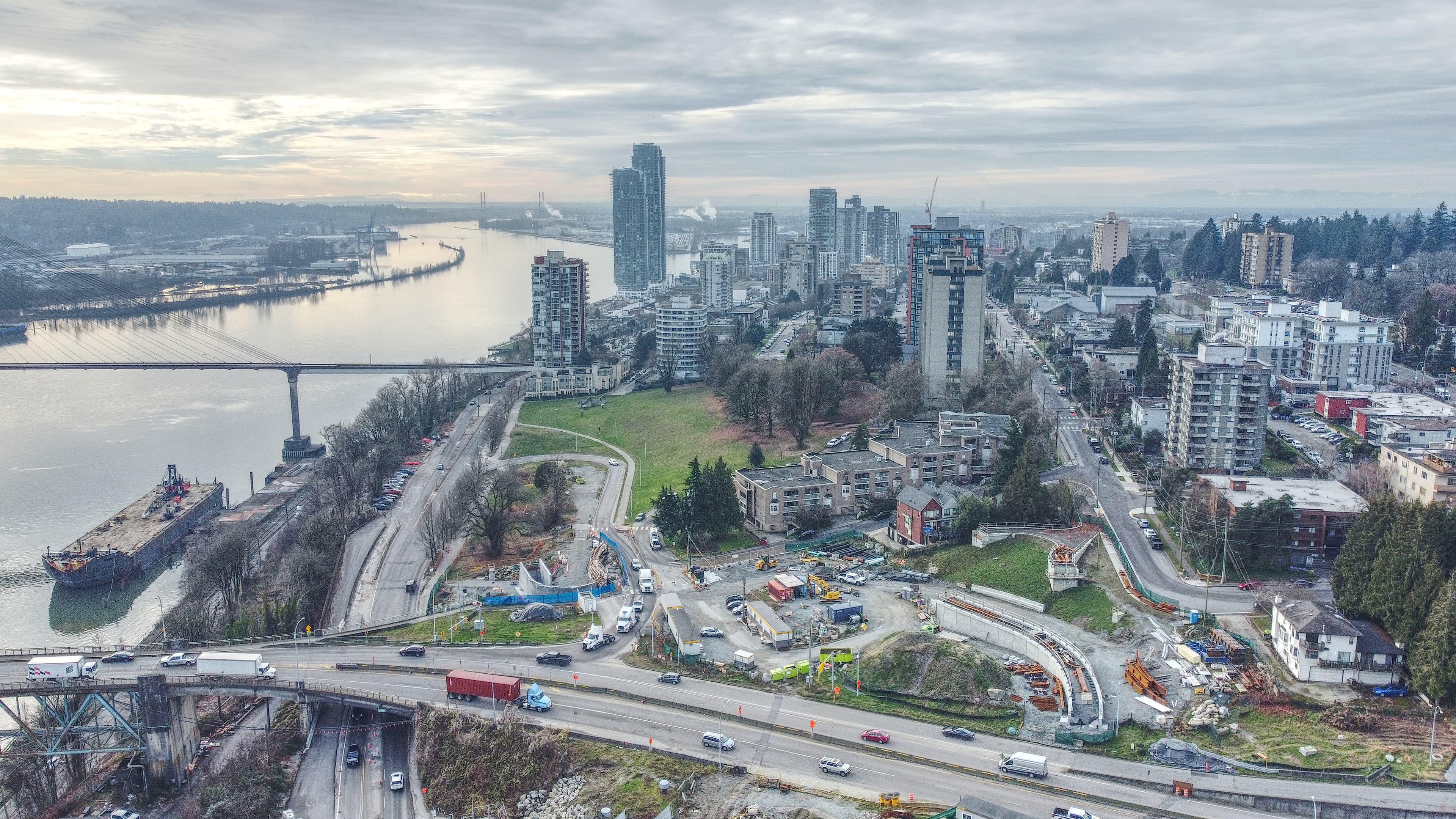 Aerial view of downtown New Westminster, British Columbia, with residential towers along the Fraser River, the Pattullo Bridge and SkyBridge crossing in the distance, and the Surrey skyline visible across the water.